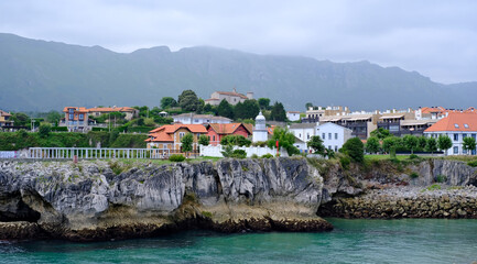 Picturesque coastal town in Northern Spain, featuring traditional houses with red roofs perched atop dramatic cliffs overlooking a turquoise bay. Llane,s, Asturias
