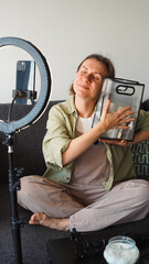 Blogger smiles into phone camera mounted on tripod and shows off cosmetics bag. Soft focus. ...