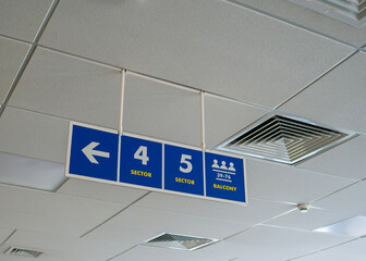 A blue sign with white arrows hanging on the ceiling in a public area indicating the balcony,...