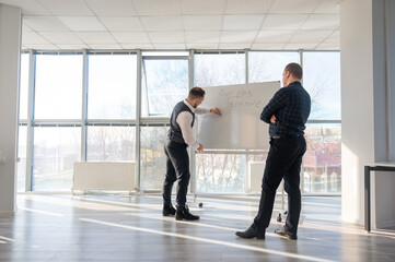 Two Caucasian men discussing work at a white board. 