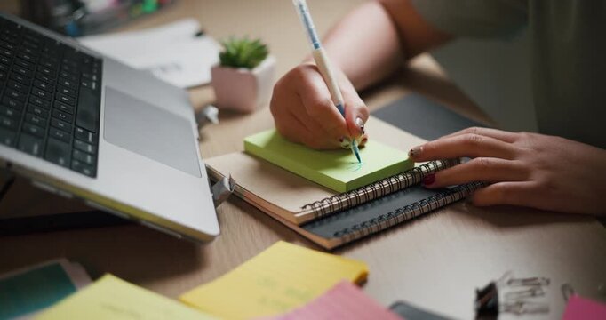 Handheld selective focus shot, Woman is writing on a green sticky note with a blue pen while sitting at a desk with a laptop and notebooks.