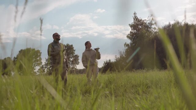 couple in matching flight suits preparing tandem glider launch over sunlit grassy field under blue sky breeze, checking harness cords, inspecting canopy inflation