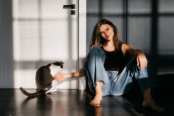 In a cozy space, a woman shares a moment of affection with a cat, their bond highlighted by play of shadows and light through large windows.