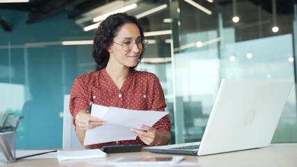 Happy young businesswoman is satisfied with results of a financial report she is reviewing sitting at a desk in office. Smiling female financier looks at documents and laptop with positive indicators