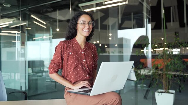 Happy young businesswoman working on laptop computer sitting at a desk in business office. Smiling female employee happily completing a task, chatting online with a colleague or browsing social media
