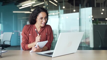 Young businesswoman watching video call conference taking notes using laptop in office. Female employee studying online, listening business training course, communicates in remote meeting with tutor - Powered by Adobe