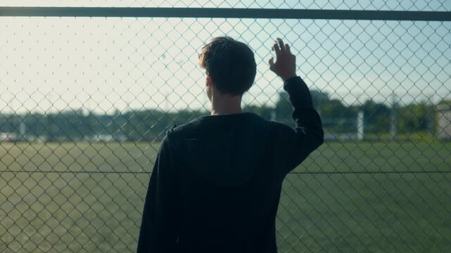Teenage boy stands facing sports fence lost in thought on sunny day