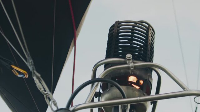 Close up of metal burner on hot air balloon with small visible flame igniting inside cylindrical structure, framed by steel pipes and wires, under bright sky during pre flight setup phase