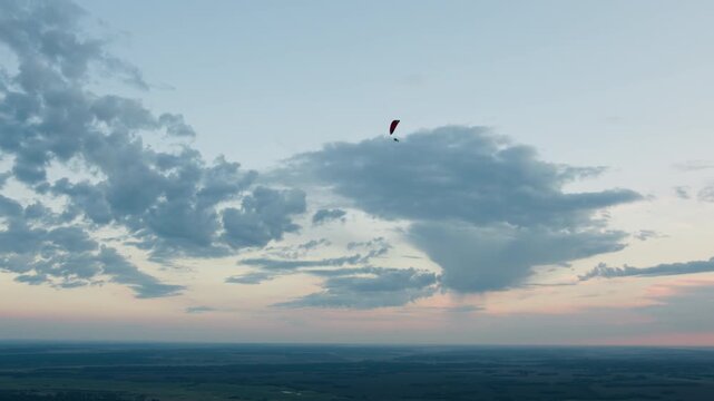 Distant silhouette of people flying powered paraglider under expansive sky with scattered clouds as they drift above farmland in gentle evening light conveying serene aerial motion