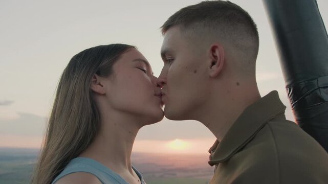 close up couples in hot air balloon floating above patchwork fields at sunset sharing intimate kiss with gentle sky tones and distant paraglider silhouette evoking romance adventure