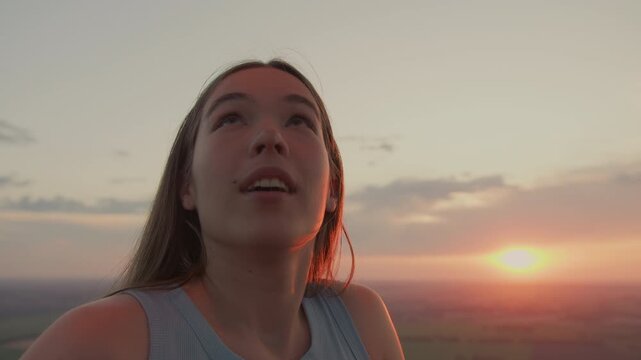 elegant young woman tilts head upward to watch metal burner flame flicker inside hot air balloon basket under warm sunset glow over sprawling fields and drifting clouds