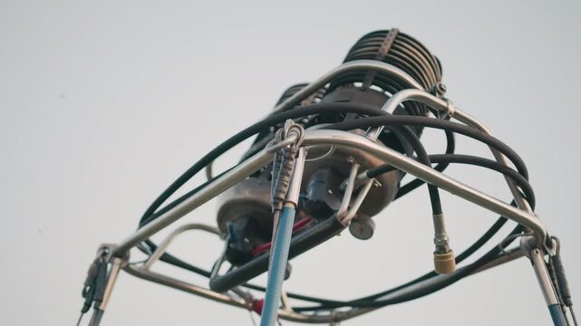 close up of hot air balloon burner assembly showing metal frame structure, braided hoses, safety cables, pressure gauge, valves and coils against clear sky