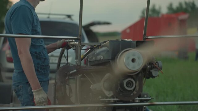 man in blue shirt and gloves stands beside spinning paramotor fan on grassy field with blurred people in background as blades rotate rapidly within metal frame