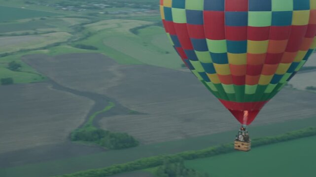 Colorful checkered hot air balloon carrying passengers drifting above patchwork field at sunset with visible flame jet, serene aerial motion over green fields and distant horizon under sky clouds