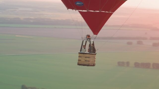 travelers in colorful hot air balloon drifting over expansive green farmland at sunset while pilot ignites burner flame adjusting altitude capturing serene sky adventure from aerial perspective