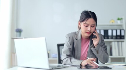 Professional Asian businesswoman working on laptop while talking on mobile phone, seated at modern office desk with corporate interior background - Powered by Adobe