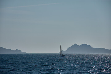 A sailboat sails at sunset through the Vigo Estuary, with the silhouettes of the Cies Islands outlined on the horizon and the shimmering waters.
