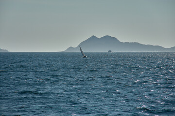 A sailboat sails at sunset through the Vigo Estuary, with the silhouettes of the Cies Islands outlined on the horizon and the shimmering waters.