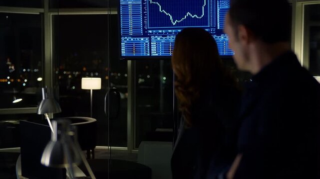 Business colleagues analyze financial data on screen at night office, discussing market trends and stock charts with concern for investment report and strategy planning - Powered by Adobe