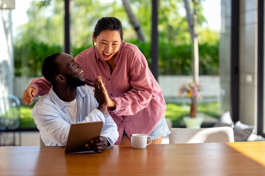 Happy young diverse, multiethnic couple planning budget, reading good news on digital tablet at home