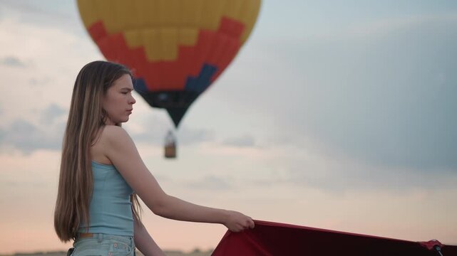 side view of young lady in denim shorts grips red hot air balloon panel as colorful envelope floats blurred in dusk sky during preflight beside rotating fan engine under pastel clouds