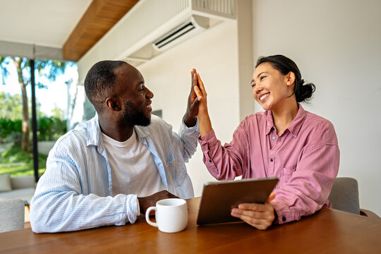 Happy young diverse, multiethnic couple planning budget, reading good news on digital tablet at home