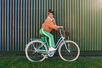 Outdoor fashion portrait of pretty african woman sitting on bike, leaning on the wall