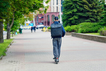 Courier with black delivery bag riding electric scooter on pavement, fast food delivery service...