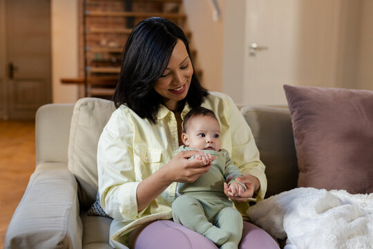 Asian mother holding infant daughter feeding with small spoon on beige couch in living room