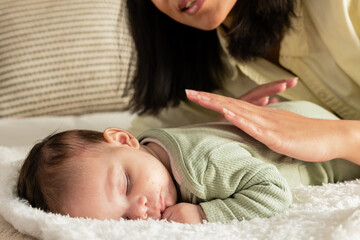 Asian mother in yellow top leaning above sleeping daughter in green onesie on plush blanket