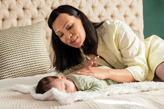 Asian mother and child leaning over infant in green onesie against tufted headboard in bedroom
