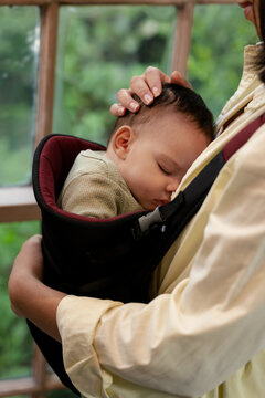 Adult caregiver and female infant cradling baby in carrier at home by wooden window frame