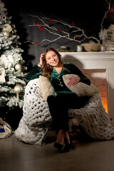 Smiling woman in a green velvet suit relaxes by a cozy Christmas fireplace and decorated tree, enjoying the festive winter season at home. Ideal for holiday and comfort themes.