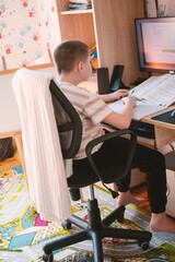 Young boy studying at a desk with a computer and book, engaged in online learning or homework in a bright room.