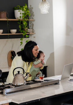 Asian mother juggling remote work while holding infant on marble kitchen island with laptop and tea