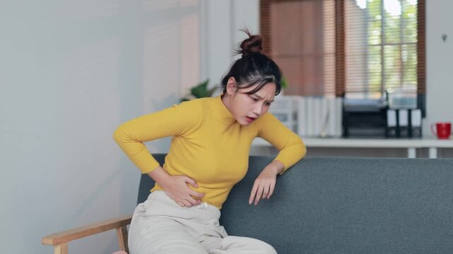Stressed young woman wincing from sharp abdominal pain while holding smartphone, experiencing sudden discomfort potentially triggered by digital communication or health issue