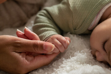 Mother gently gripping infant daughter's hand on soft white plush blanket in nursery