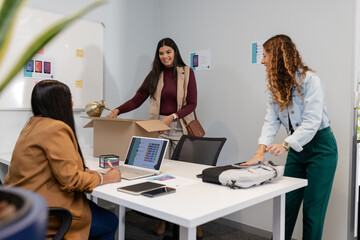 Diverse female colleagues unpacking desk lamp box and sorting backpack at office table with laptop