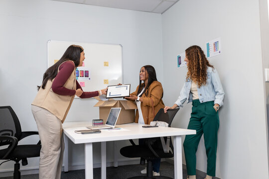 Diverse female colleagues exchanging tablet while reviewing laptop, notes and whiteboard in office