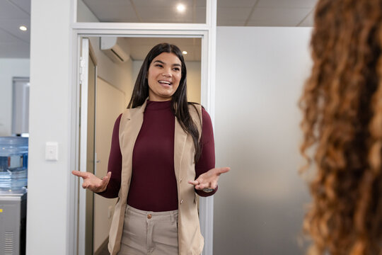 Diverse female coworkers standing in office hallway wearing blazer gesturing near water cooler