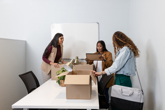 Diverse female coworkers unpacking desk lamp and potted plant from cardboard box in small office