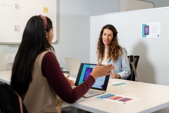 Diverse female colleagues sitting at office table reviewing UI design with laptops and color swatch