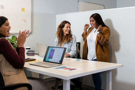 Diverse female team clapping, smiling, raising fists around laptops in office meeting room