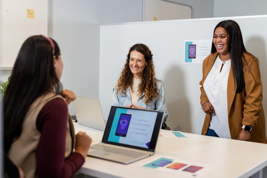 Diverse female team collaborating at meeting table reviewing app prototype on laptops with mockups