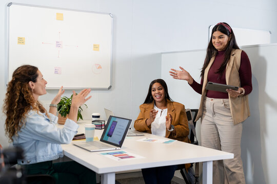 Diverse female coworkers collaborating at office table using laptops tablet charts coffee cup plant
