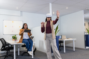 Diverse female coworkers using VR headset and tapping tablet in open office with laptops, plants