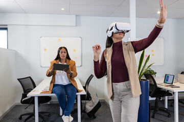 Diverse female coworkers using VR headset gesturing toward whiteboards at office with tablet