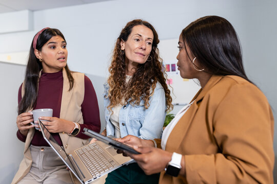 Diverse female colleagues gathering in bright office workspace with laptop tablet sticky notes
