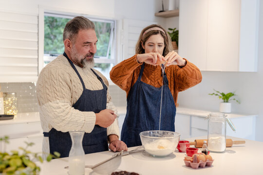 Couple wearing blue aprons cooking at kitchen island cracking egg into bowl while holding whisk