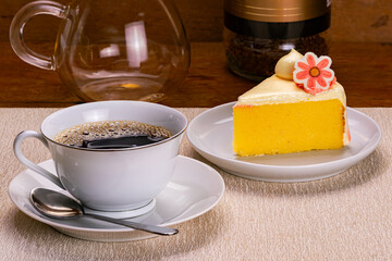 Side view of a cup of black coffee in white ceramic cup with a metal spoon on white ceramic saucer and slice of vanilla sponge cake garnished with cream in white ceramic dish on light brown table mat.